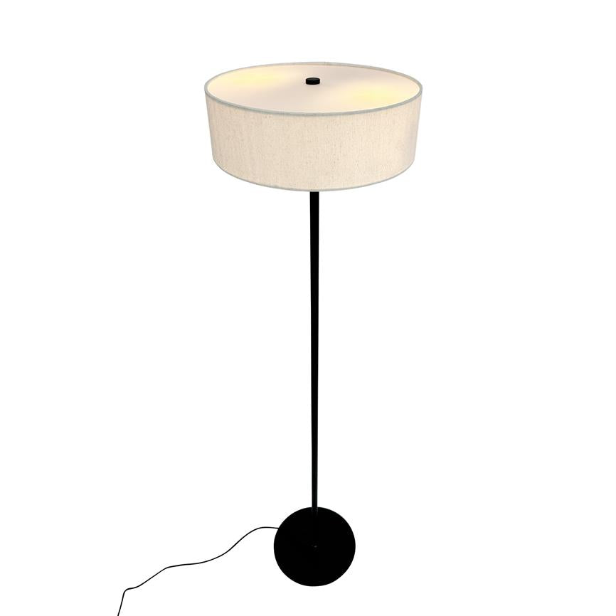 En stålampe udsender blødt, varmt lys i en enkel hvid baggrund; den har en høj, slank sort stang, rund sort fod og en stor beige cylindrisk lampeskærm.