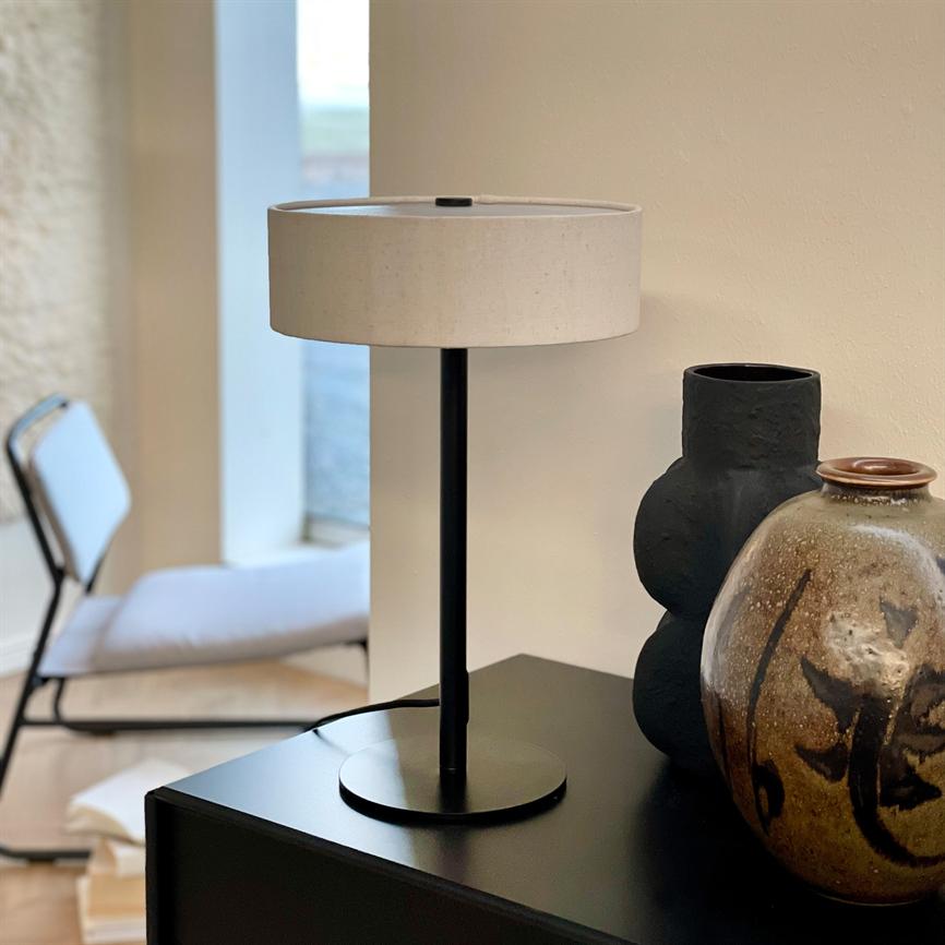 En bordlampe med sort metalfod og hvid skærm står på et sort skab ved siden af to keramiske vaser; i baggrunden ses en stol og vindue i lyst rum.