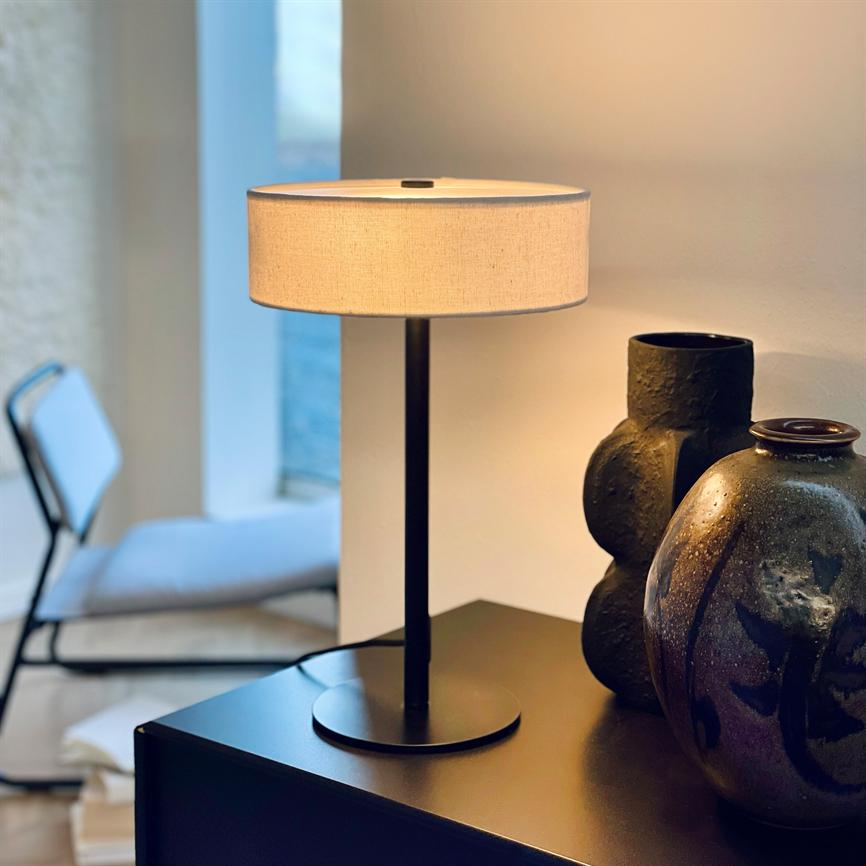 Bordlampe står tændt på et sort skab, kaster varmt lys over to mørke, skulpturelle keramikvaser; i baggrunden en enkel stol og et lyst vindue i et roligt rum.