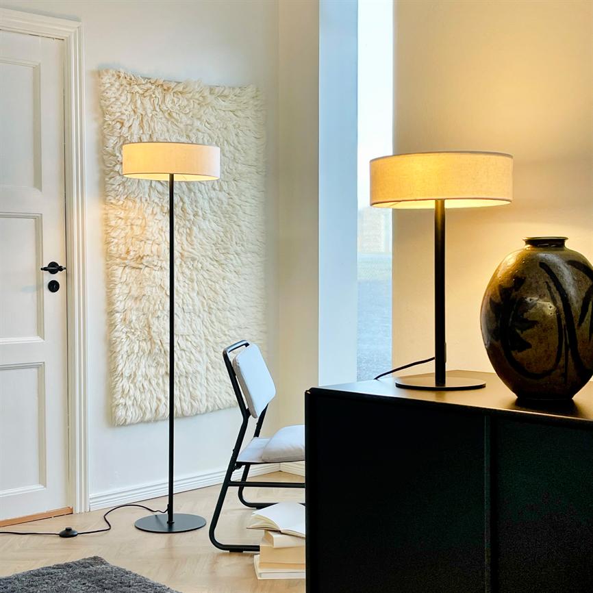Gulvlampe lyser blødt i et lyst hjørne; en matchende bordlampe står tændt på sort skænk ved en stor keramisk vase, hvid stol og uldtæppe på væggen.