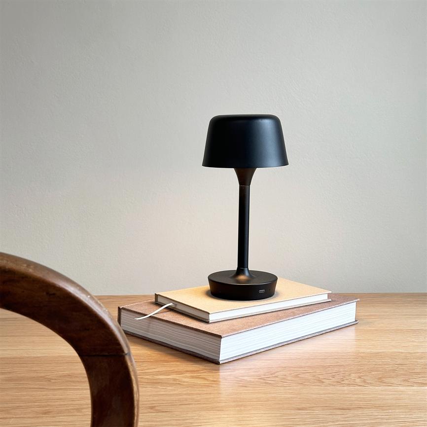 Sort bordlampe står og udsender blødt lys på to stablerede bøger på et lyst træbord foran en neutral væg; en buet træstol ses i forgrunden.