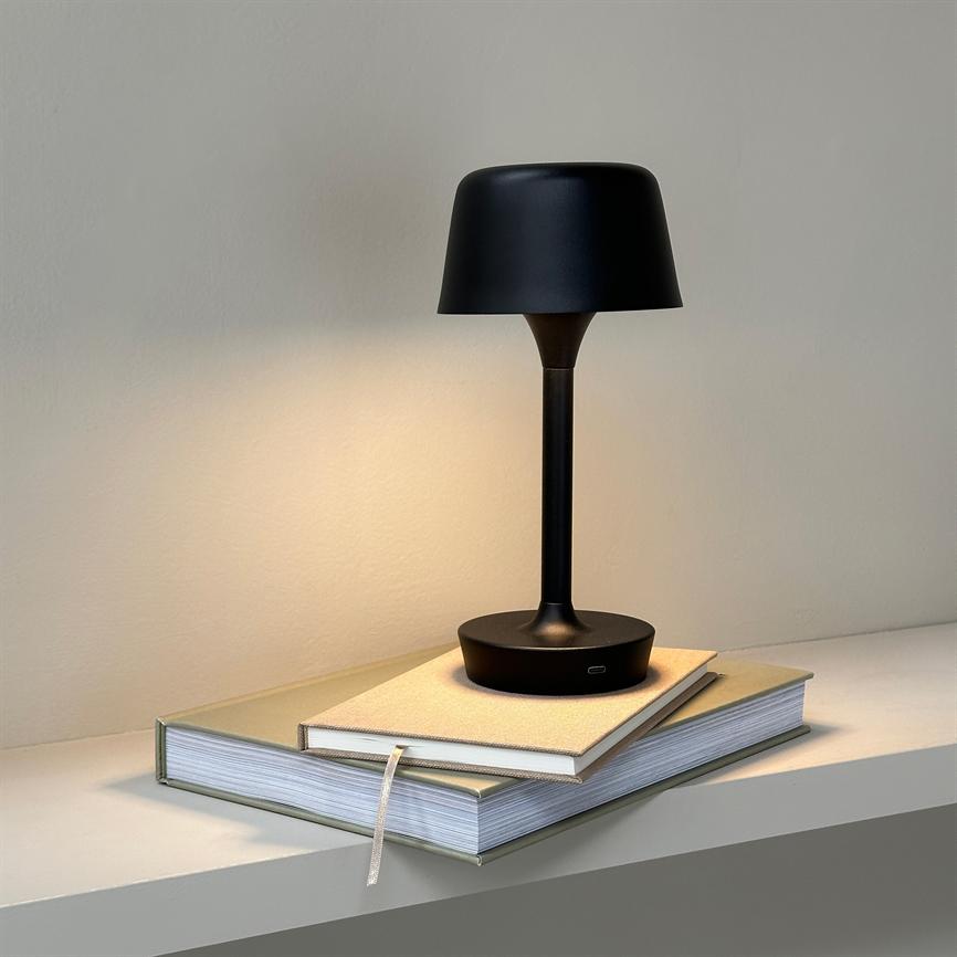 Sort, moderne bordlampe lyser blødt nedad og står på to stablede bøger med et båndbogmærke, placeret på en enkel hylde foran en neutral, lys væg.