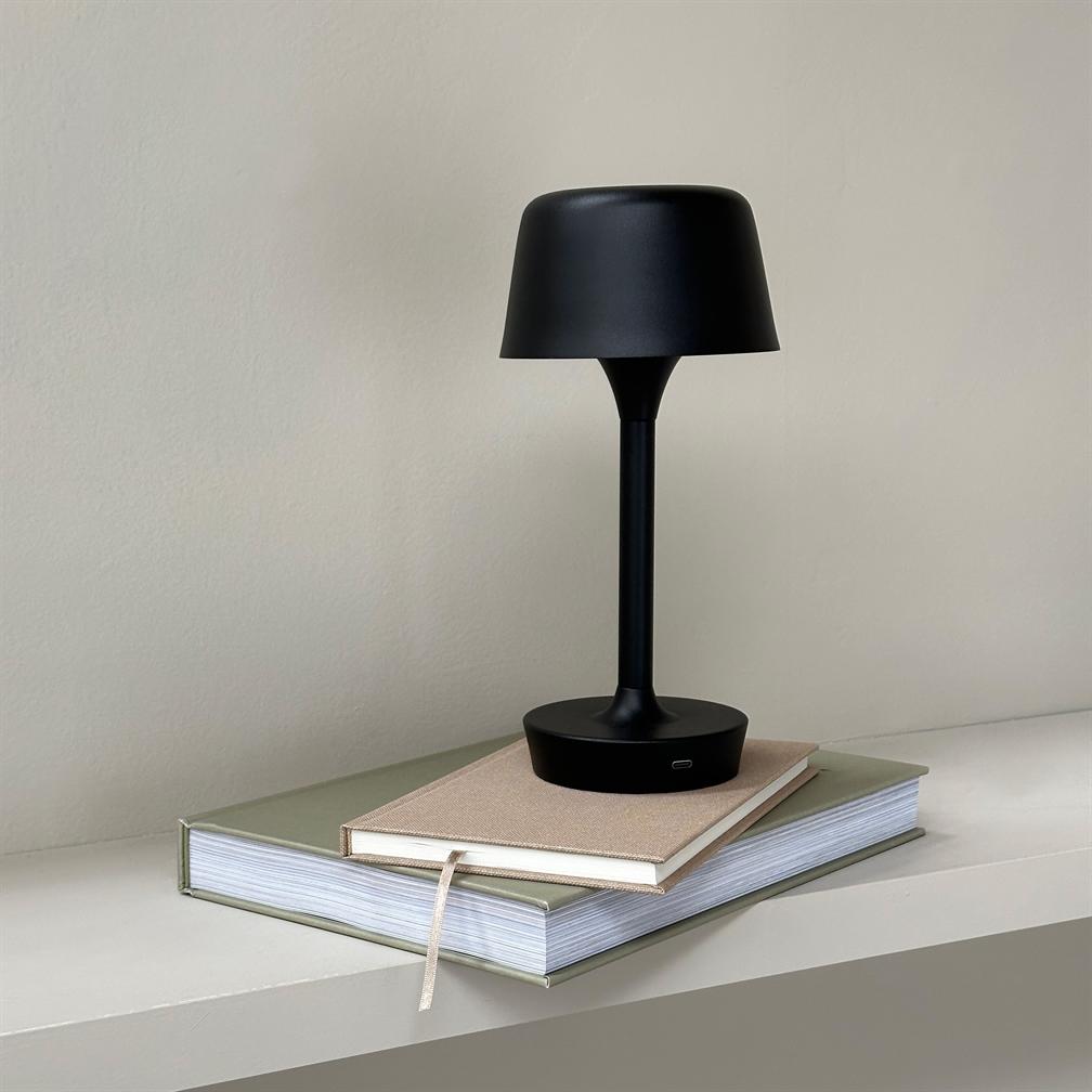 Sort bordlampe står på to stablede bøger på en lys hylde; mat sort skærm og slank fod, neutral beige væg i baggrunden.