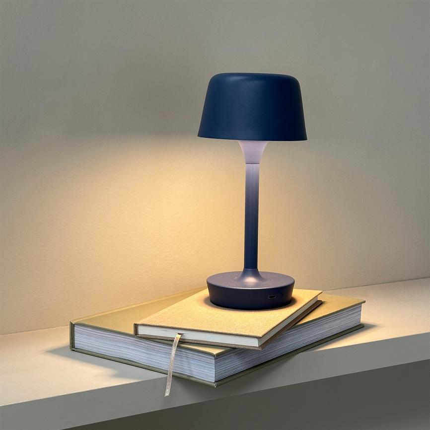 En blå bordlampe står tændt på to stakkede notesbøger; den kaster varmt lys på en neutral væg og en enkel hylde i et roligt indendørs miljø.