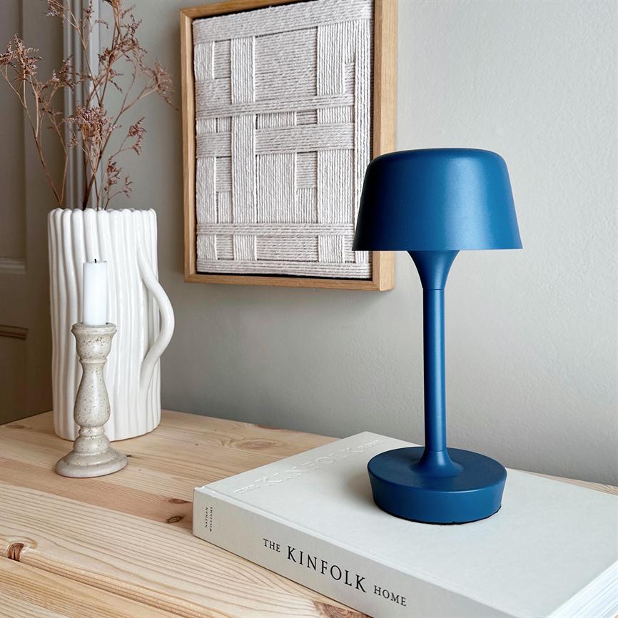 Blå bordlampe står på en bog, omgivet af en lys trækommode med hvid vase, lysestage og indrammet vævet kunst på en neutral væg. Tekst: "THE KINFOLK HOME".