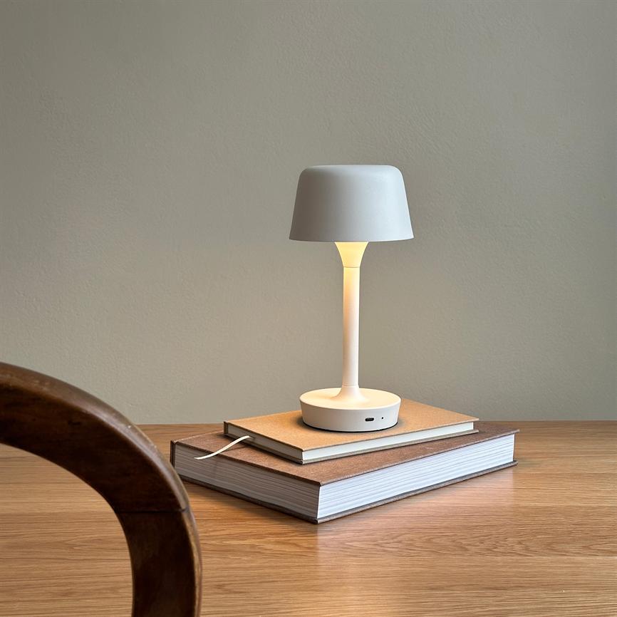 Hvid bordlampe lyser blødt; står på en stak bøger på et træbord i et roligt rum med lyse vægge. En rund trækant af en stol ses i forgrunden.