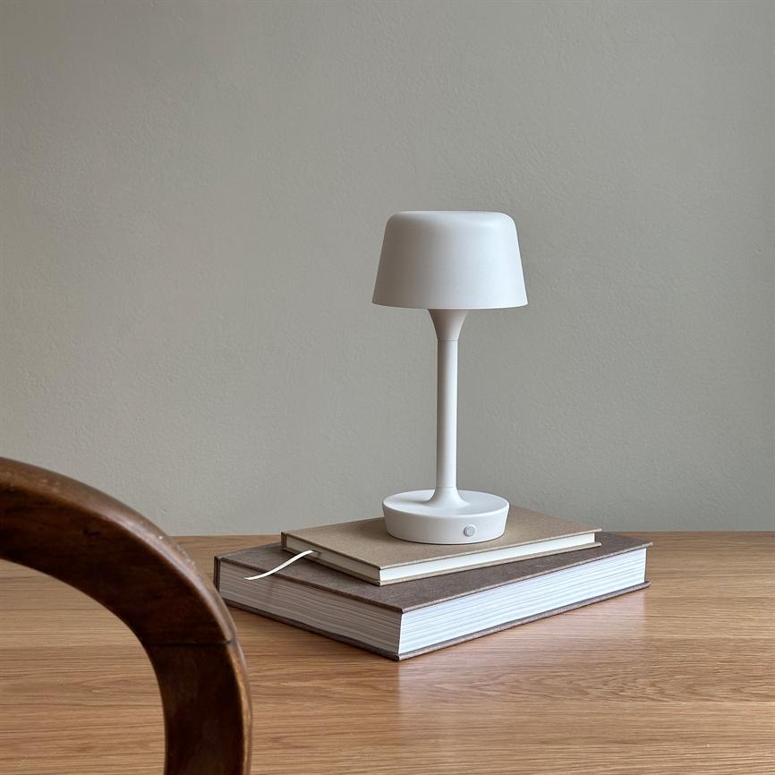 Bordlampe står på to stakede bøger på et træbord; den hvide lampe er slank og enkel, med en buet lampskærm, mod en lysegrå væg, del af en træstol i forgrunden.