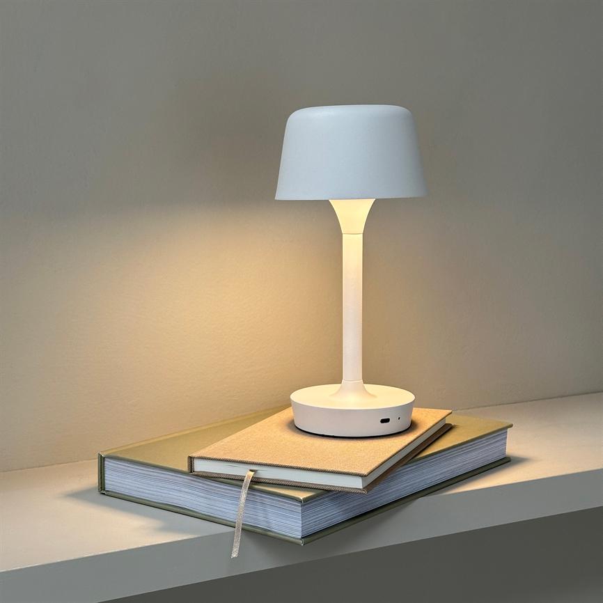 Hvid bordlampe kaster blødt, varmt lys; den står på to stablede bøger på en hylde foran en neutral, lys væg.