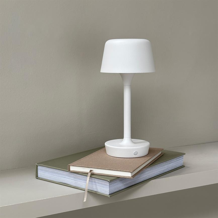 Hvid bordlampe hviler på en beige notesbog oven på en tyk grøn bog, på en hylde foran en lysgrå væg.