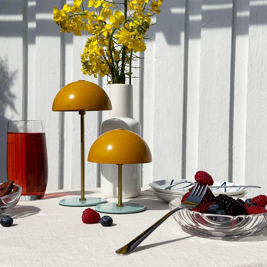 To gule kuppel-lamper står på en hvid dug, foran en hvid vase med gule blomster; bær i glasskål, en gaffel og et glas rød drik ligger på et solbelyst udendørs bord.