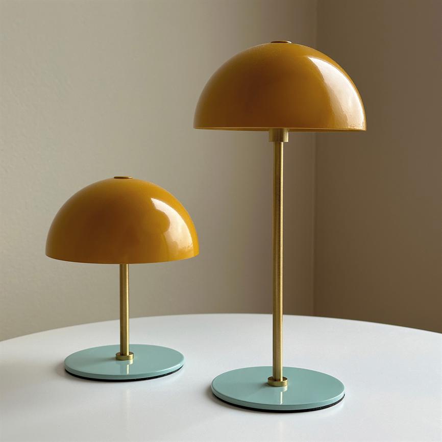To gule, kuppelformede lamper står på et rundt hvidt bord; den ene højere end den anden, med gyldne stængler og mintgrønne runde sokler foran en beige væg.