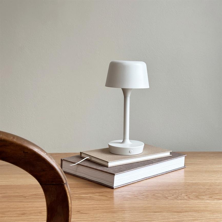 En hvid bordlampe står hvilende på to stablede bøger på et lyst trækøkkenbord, med en buet træstol i forgrunden og en neutral, beige væg i baggrunden.