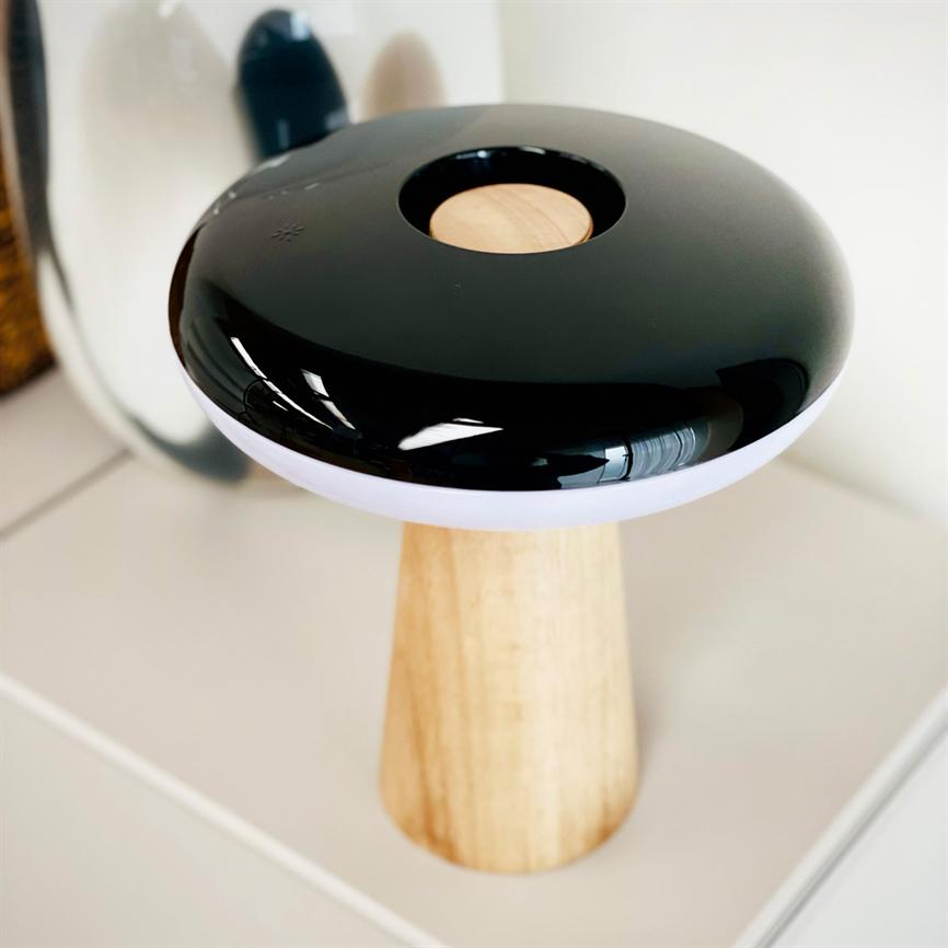 Sort, blank, svampelignende lampe med rund træindsats og hvid kant står på en lys træfod på en hvid bordplade og reflekteres i et spejl bagved.