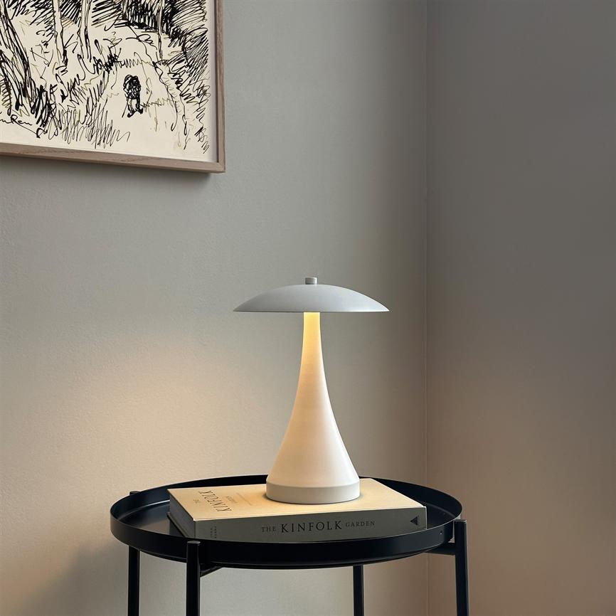 En hvid, svampelignende bordlampe udsender blødt, varmt lys; den står på en bog på et rundt sort sidebord i et roligt hjørne under en indrammet tegning på en grå væg.
 
 Tekst i billedet: "THE KINFOLK GARDEN"