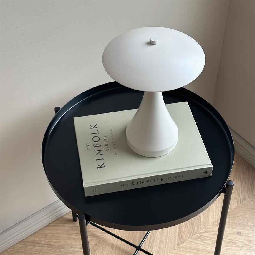 En lille hvid bordlampe står på en lukket bog på et rundt sort sidebord i et lyst hjørne ved trægulv og væg. Tekst på bogen: "THE KINFOLK" "GARDEN" (ryg: "THE KINFOLK GARDEN").