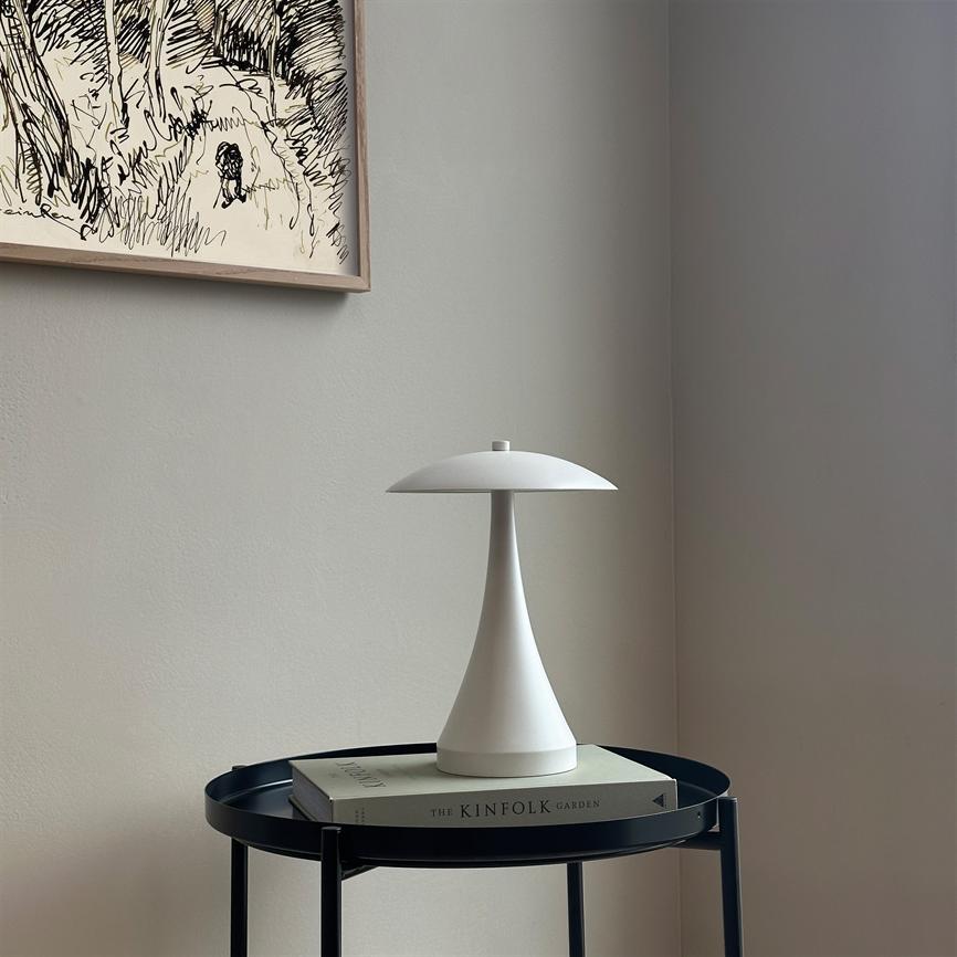 En hvid, svampelignende bordlampe står på en bog på et rundt, sort sidebord i et lyst, minimalistisk hjørne. Tekst på bogryg: "THE KINFOLK GARDEN".