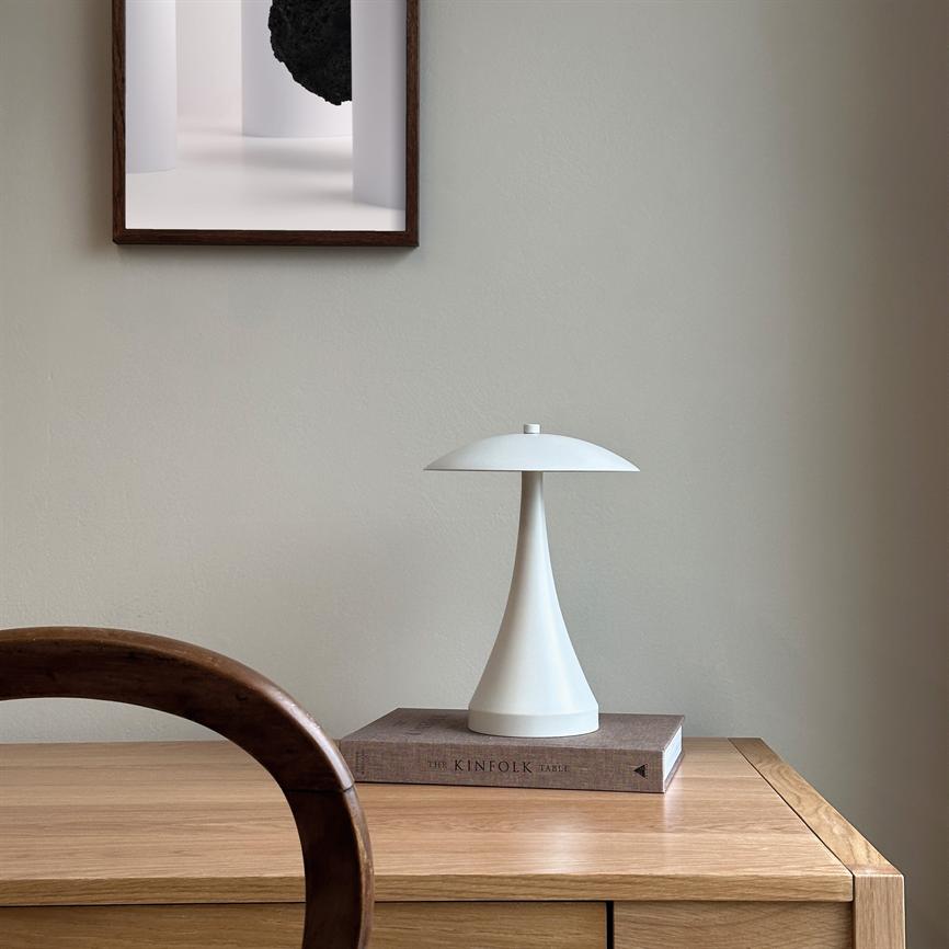 Hvid bordlampe står på en bog ovenpå et lyst træskrivbord; rund træstol i forgrunden, neutral væg og indrammet kunst hænger over. Tekst på bog: THE KINFOLK TABLE.