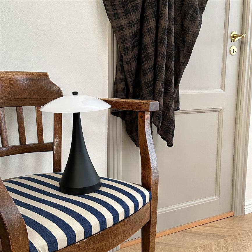 Sort konisk bordlampe med hvid skærm står på en træstol. Omkring ses et blåt-hvidt stribet sæde, en ternet jakke hængende på døren og lyst trægulv.