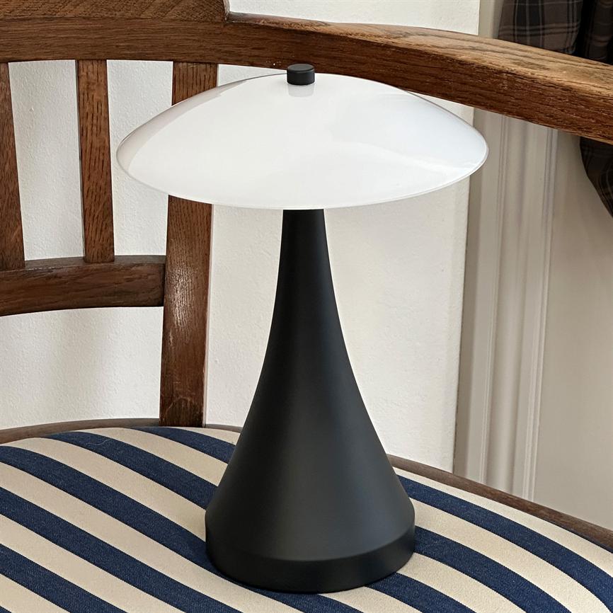 Bordlampe med mat sort, kegleformet fod og rund hvid skærm står på en blå- og hvidstribet polstret træstol foran en hvid væg.