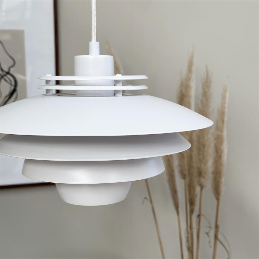 En hvid, lagdelt pendel lampe hænger i en tekstilklædt ledning; den pryder et roligt interiør foran en beige væg med tørrede pampasgræs og et indrammet abstrakt print.
