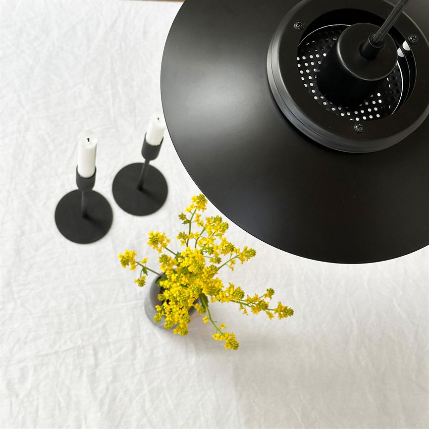 Sort loftslampe hænger over en hvid dug, oplyser en lille vase med gule blomster; to minimalistiske sorte lysestager med hvide stearinlys står på dugens overflade.