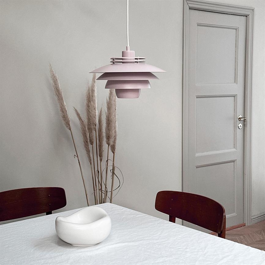 En lyserød, lagdelt loftlampe hænger over et hvidt duget spisebord; en hvid keramikskål står på bordet, omgivet af tørrede pampasstrå, mørke træstole og en grå dør bagved.