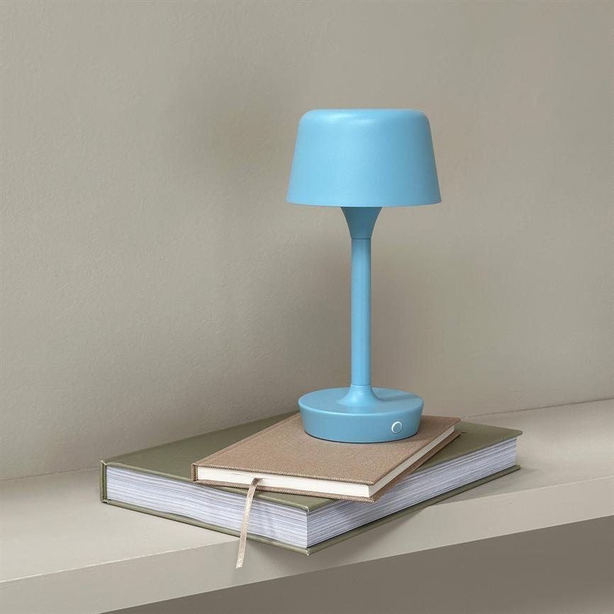 En blå bordlampe står slukket på to stablerede bøger på en smal, lys hylde foran en neutral beige væg.