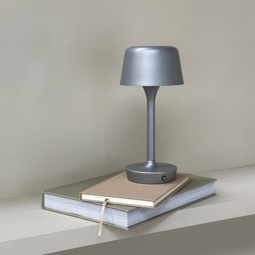 En mat sølvgrå bordlampe står slukket på to stablede notesbøger med et synligt bogmærke, på en hylde foran en lys beige væg.