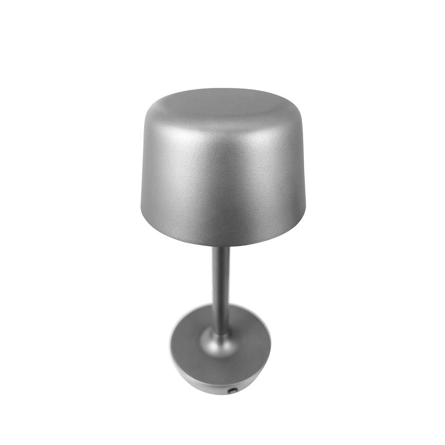 Sølvfarvet bordlampe står oprejst på hvid baggrund; et slankt skaft forbinder en afrundet cylindrisk skærm og en rund base, mat metalfinish, fotograferet skråt oppefra.