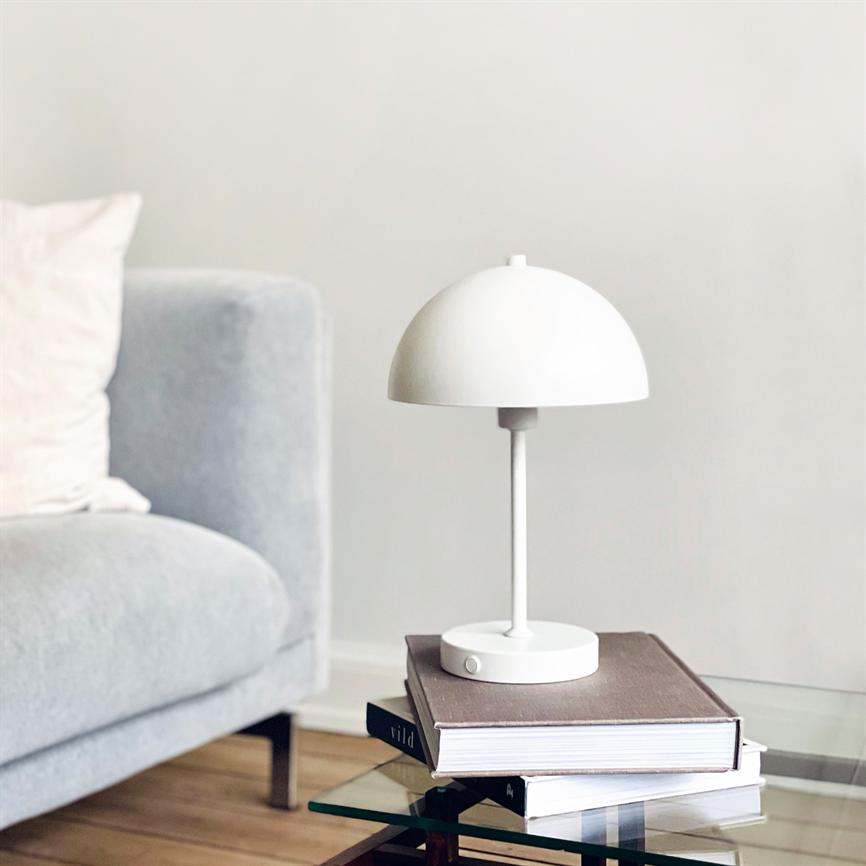 Hvid kuppelformet bordlampe står på en bunke bøger; skærmen kaster blødt lys over et glas-sofabord; grå sofa ses i baggrunden. Tekst på bogryg: "vild".