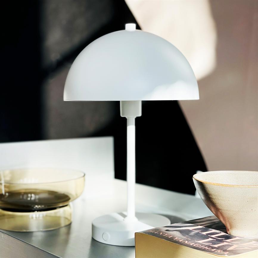 En hvid, kuppelformet bordlampe står på et bord; ved siden af to gennemsigtige glasskåle, en keramisk skål og en bog på en lys overflade foran en mørk baggrund.