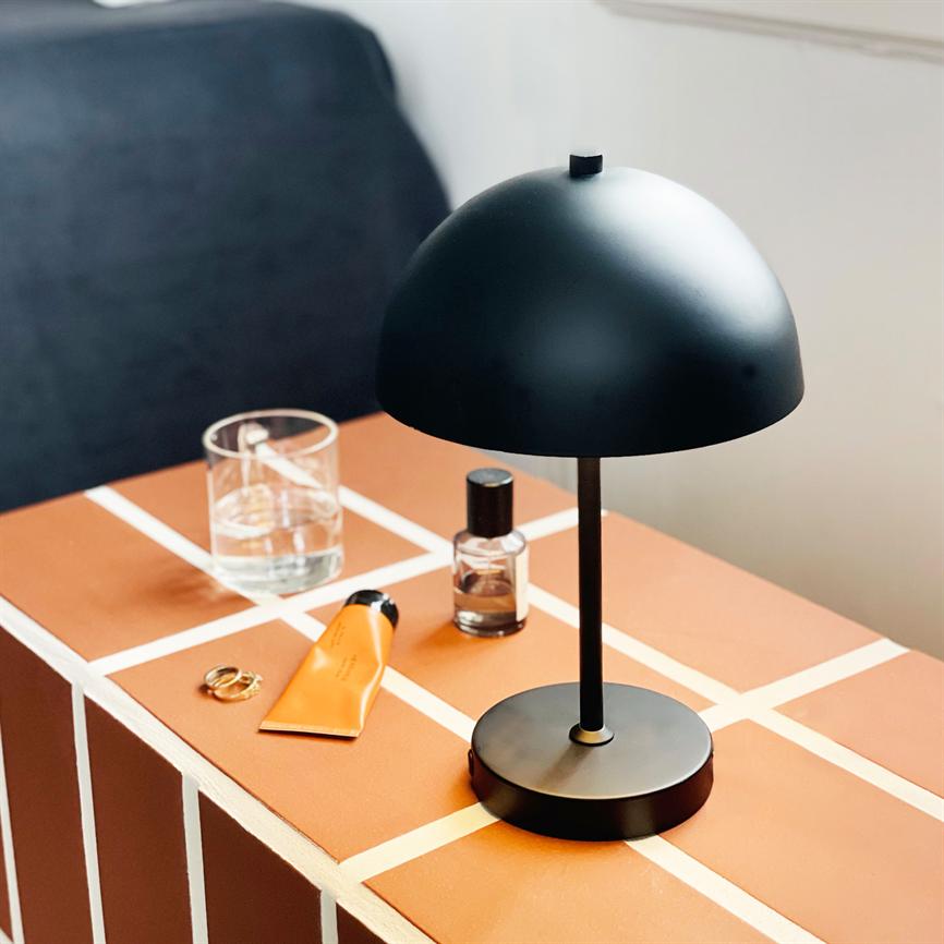 Sort bordlampe står på et orange flise-mønstret sidebord; ved siden af et glas vand, en parfume, en orange tube og en ring, foran en mørk sofa og en hvid væg.