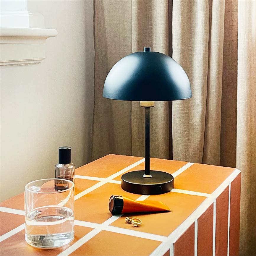 Sort kuppellampe står på et orange bord med hvide fuger; ved siden af ligger et glas vand, en orange tube og en guldring, foran lyse gardiner og en vindueskarm.