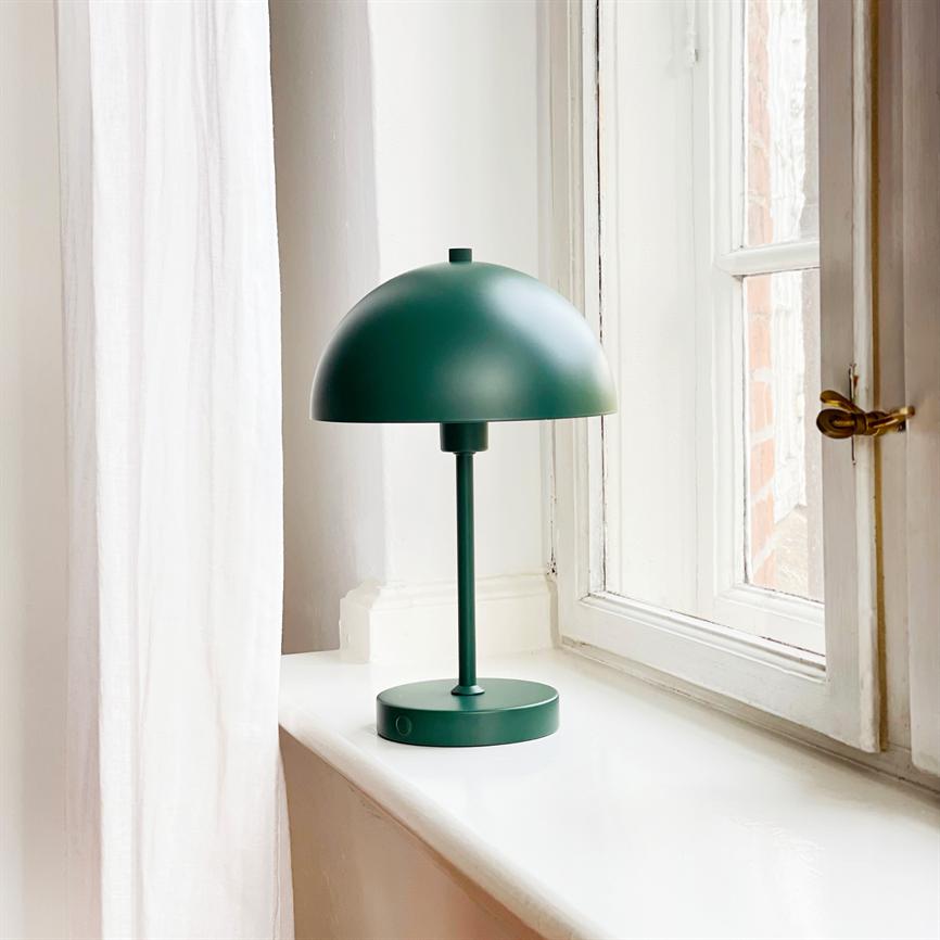 Grøn metalbordlampe står på en hvid vindueskarm ved et vindue; blødt dagslys filtrerer ind, foran et lyst gardin og et åbent sprossevindue.