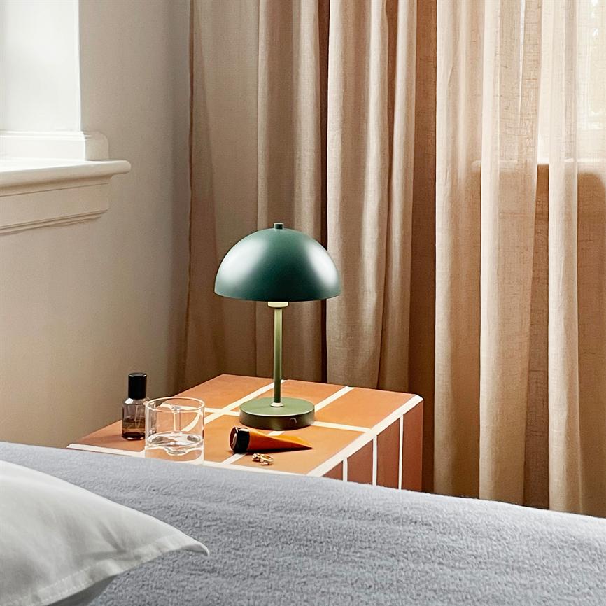 En grøn kuppelbordlampe står på et orange ternet natbord, omgivet af et glas, en parfumeflaske og nøgler, ved en seng foran beige gardiner i et roligt soveværelse.