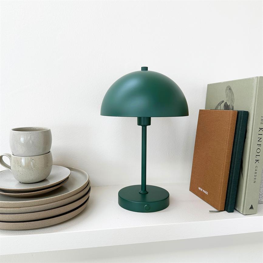 En mørkegrøn bordlampe står slukket på en hvid hylde, flankeret af beige tallerkener og kopper til venstre og bøger til højre. Tekst: "THE KINFOLK GARDEN"; "NEW MAGS".