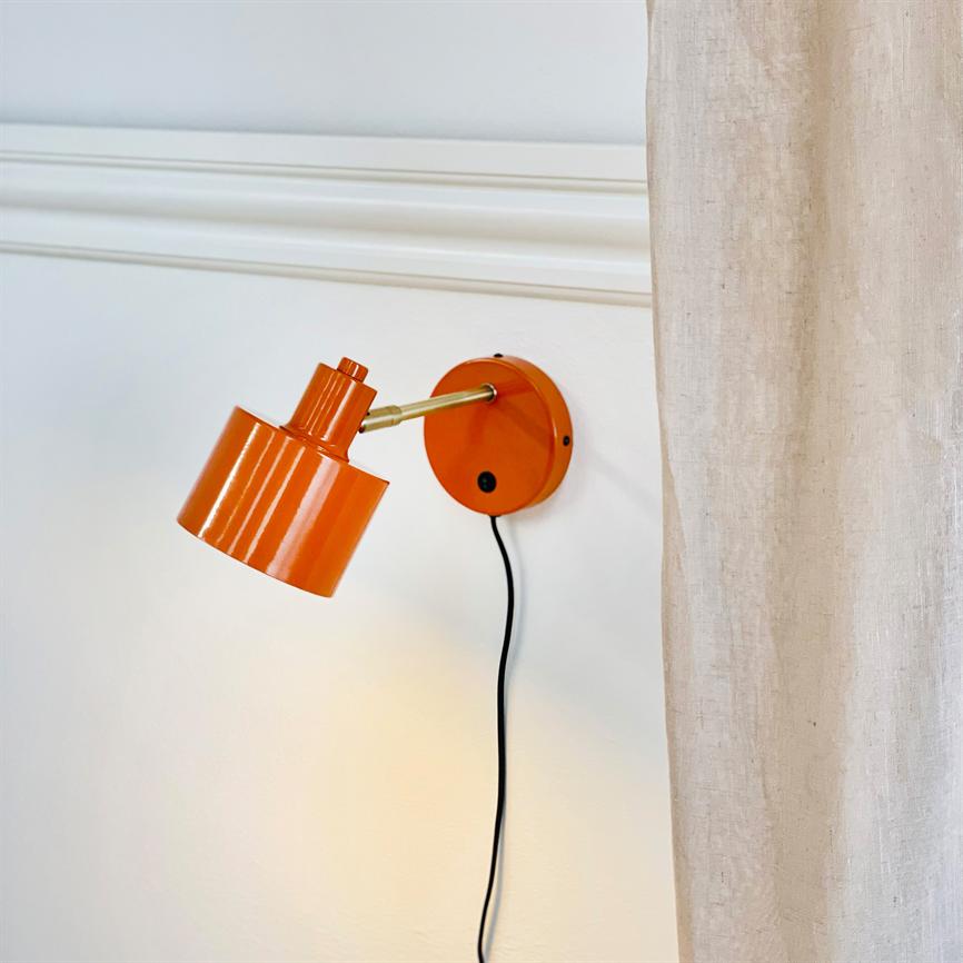 Orange metal væglampe monteret på en messingarm peger skråt nedad og kaster varmt lys på en hvid væg; en sort ledning hænger ned ved siden af et lyst naturfarvet gardin.