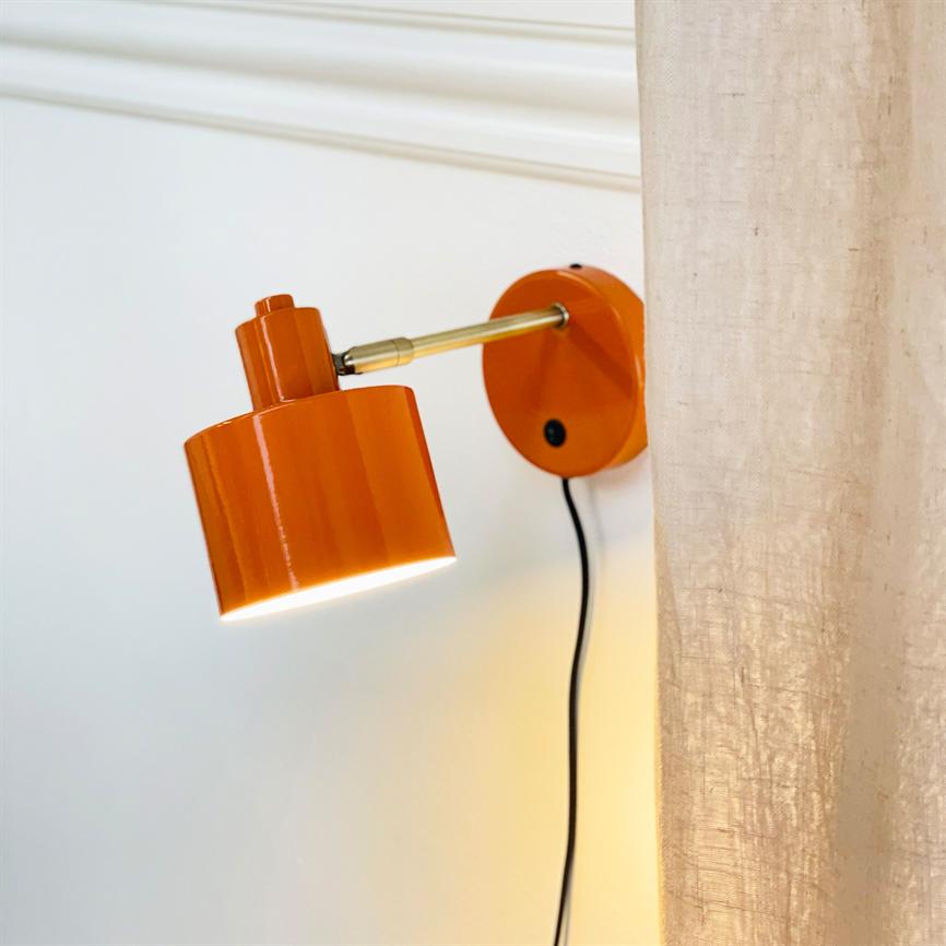 Orange væglampe lyser nedad fra en messingarm, monteret på en hvid væg med sort ledning; til højre hænger et lyst, gennemsigtigt gardin.