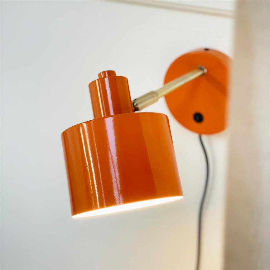 Orange væglampe, tændt og kaster lys nedad; monteret på væg med messingarm og sort ledning, foran en lys hvid væg og synlig gardinkant.