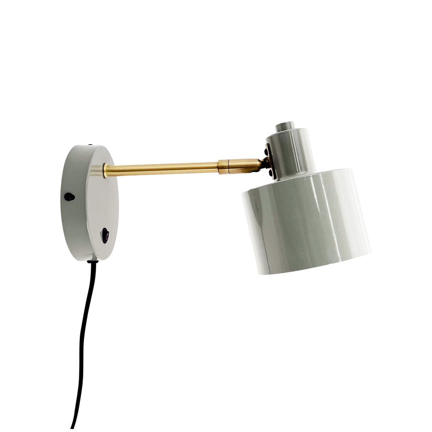 Væglampe med rund vægbeslag og messingarm, justerbar hvid cylindrisk skærm peger skråt nedad; sort ledning hænger ned fra beslaget mod en ren hvid baggrund.