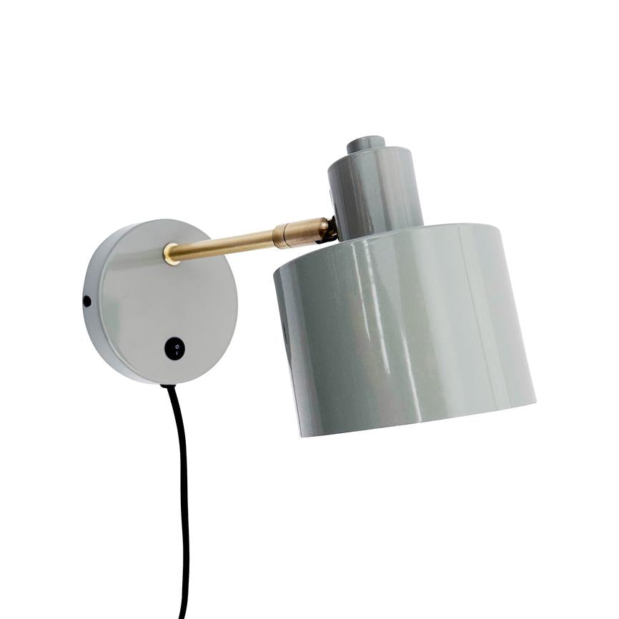 Væglampe monteret på rund vægplade, en messingarm holder et justerbart gråt metal lampehoved, lampen peger nedad og har et sort hængende strømkabel; hvid baggrund.