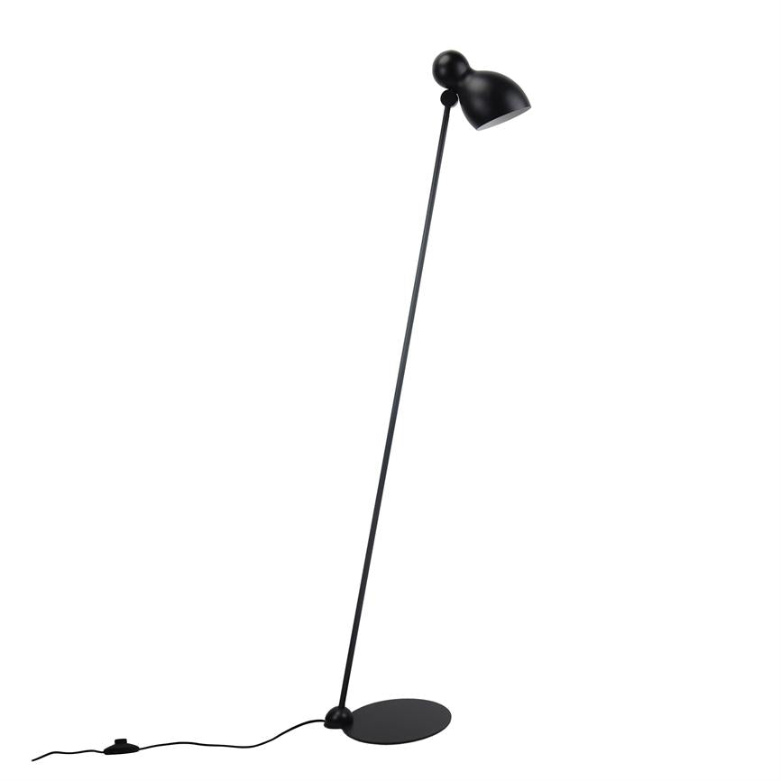 Sort gulvlampe står skråt med afrundet skærm og tynd stang på rund bundplade; synlig ledning med fodkontakt på hvid baggrund.