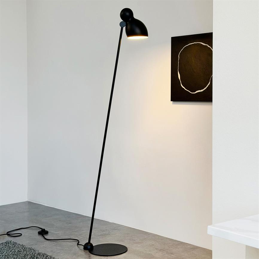En slank sort gulvlampe lyser nedad og står skråt på betongulv foran en hvid væg; et lille sort maleri med en hvid cirkel hænger delvist til højre.