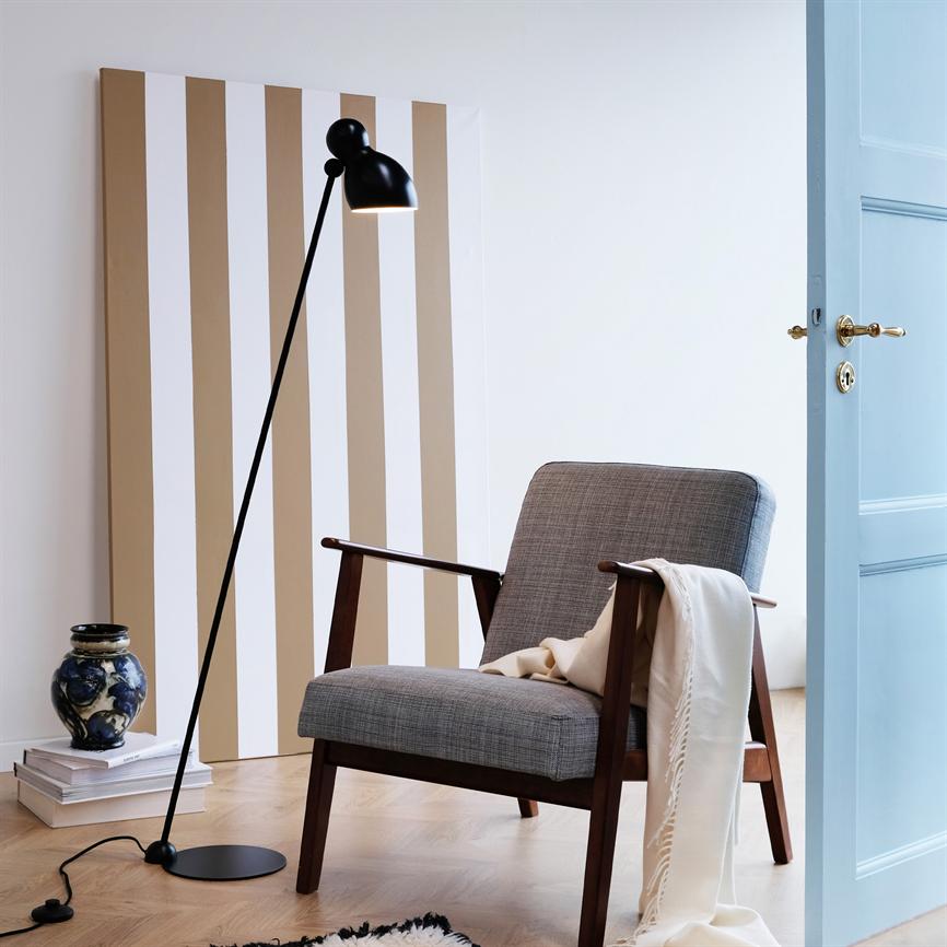 En grå polstret lænestol med træstel har et lyst tæppe over armlænet; den står ved en sort gulvlampe foran et beige-hvid stribet lærred, tæt på en åben lyseblå dør.