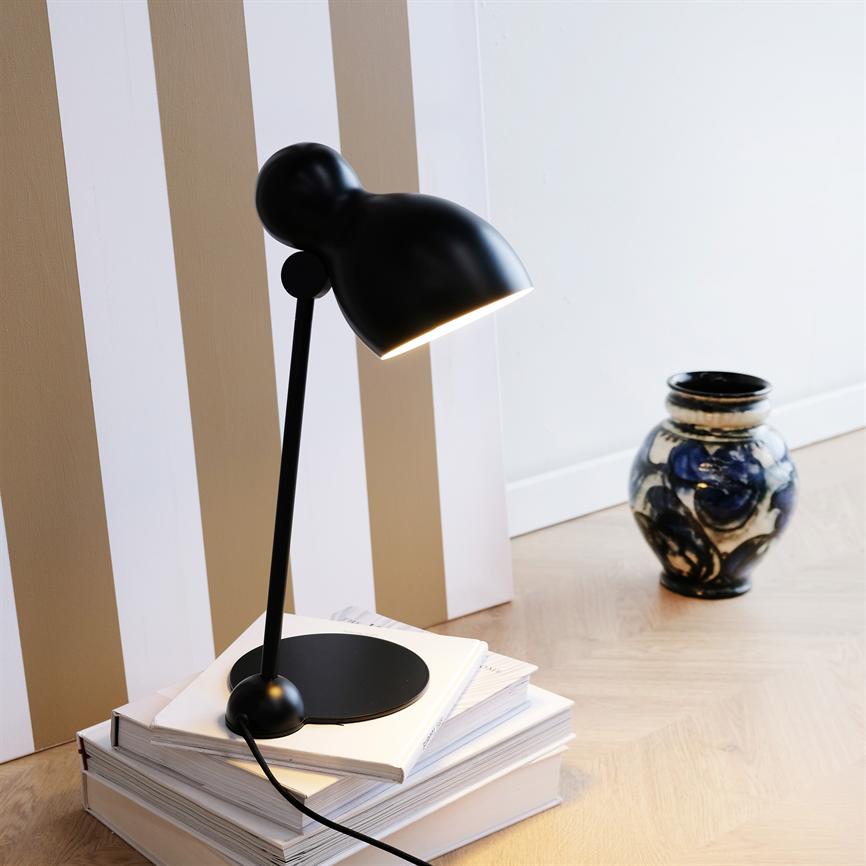 Sort bordlampe lyser ned på en stak hvide bøger på trægulvet, foran en beige-hvid stribet væg; i baggrunden står en blå-hvid keramisk vase.