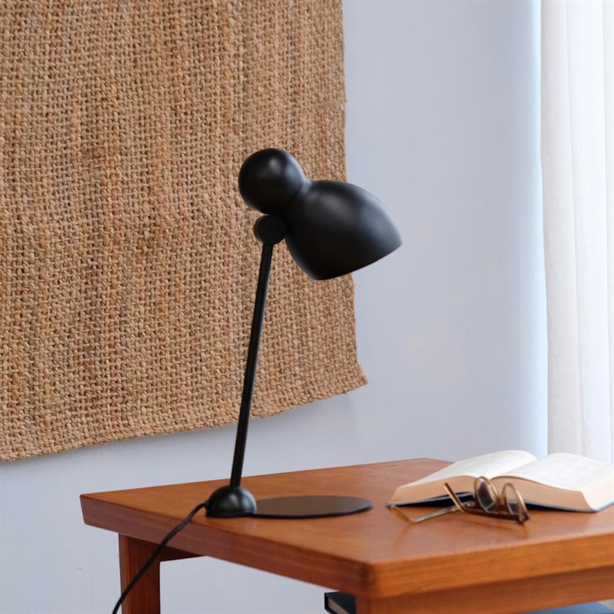 Sort skrivebordslampe vipper nedad over et træbord; en åben bog og et par briller ligger ved siden af. Bagved hænger et beige vævet tæppe, til højre hvide gardiner.