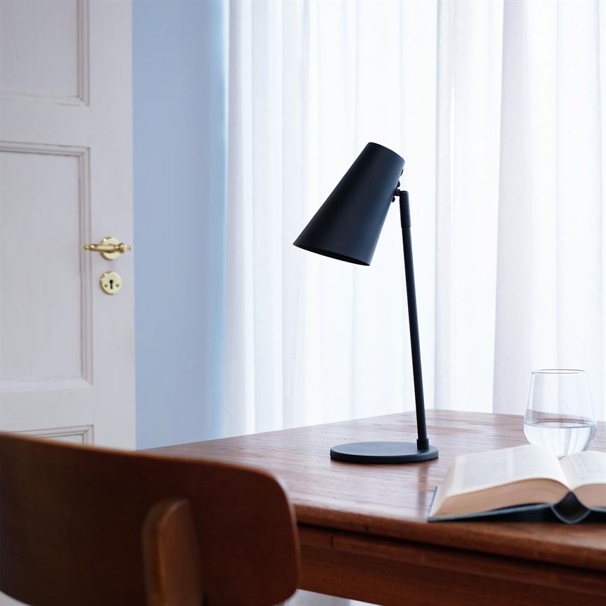 En sort bordlampe står med skærmen vinklet nedad på et træbord, omgivet af en åben bog, et glas vand, en stol og lyse gardiner foran en dør.