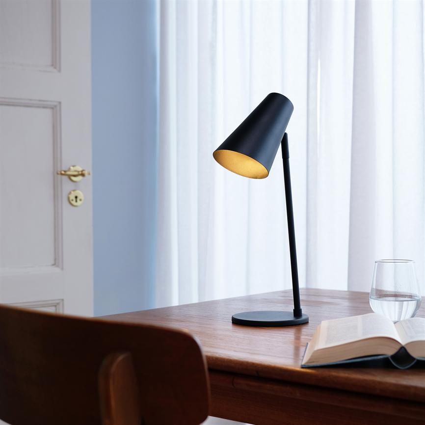 En sort bordlampe lyser blødt over et træskrivebord; en åben bog og et glas vand står ved siden af, med en stol forrest og lyse gardiner og en dør bagved.