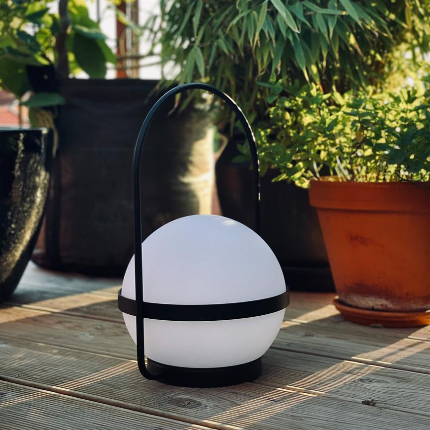 En rund, hvid bærbar lampe hviler på en træterrasse; dens buede sorte håndtag står op, omgivet af lerpotter med grønne planter i varmt, skråt sollys.