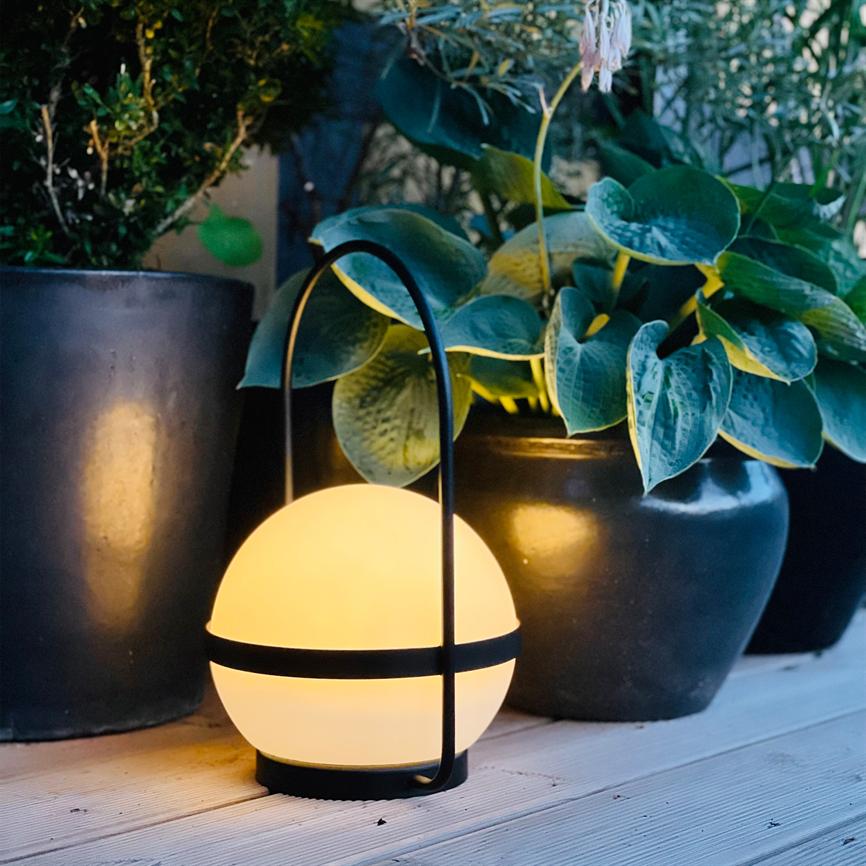 En kugleformet lampe lyser varmt; den står på en træterrasse mellem store, sorte urtepotter. Grønne, brede bladplanter danner baggrund og kaster bløde skygger.